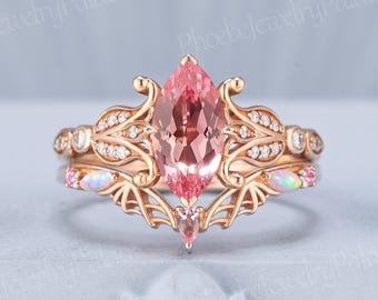 May include: A rose gold engagement ring set featuring a marquise-cut pink gemstone as the centerpiece. The ring is adorned with smaller white gemstones and intricate filigree detailing. A matching band with opal accents completes the set, showcasing a delicate and elegant design.