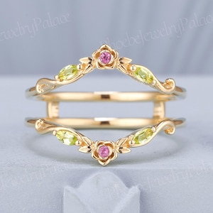 May include: A gold-colored ring guard with floral and leaf accents. The ring features pink and green gemstones. The design includes two curved bands with decorative elements, creating a unique and elegant look. The ring is set against a light background.