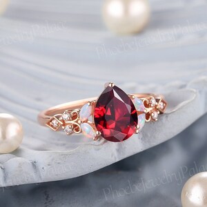 May include: A pear-shaped red gemstone ring with a rose gold band. The ring is set with smaller white gemstones and has a delicate floral design.
