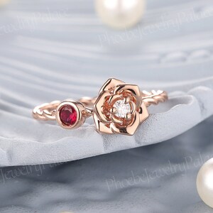 May include: A rose gold ring with a white diamond and a red ruby. The ring features a rose design with a twisted band.