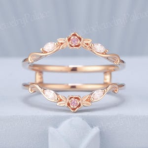 May include: Rose gold ring guard with floral and leaf accents. The ring features a small pink gemstone in the center of each flower and marquise-cut white gemstones. The ring is designed to be worn with another ring.