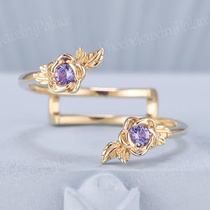 May include: A gold-colored ring with a floral design. Each flower features a purple gemstone center and is accompanied by gold-colored leaves. The ring's band has a unique, open design. The ring is displayed on a light gray surface.