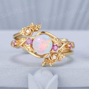 May include: A gold-colored ring set featuring an opal gemstone at the center, surrounded by smaller pink gemstones and floral accents. The ring has a delicate, nature-inspired design with leaves and small diamonds.