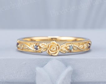Op de afbeelding: Een gouden ring met een centraal gebeeldhouwd rozenmotief, geflankeerd door ingewikkelde bladgravures en kleine, ronde, donkergekleurde edelstenen die langs de band zijn gezet. Het metalen oppervlak heeft een getextureerde afwerking met kralenranden.