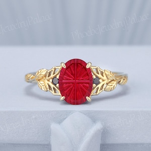 May include: A gold ring featuring an oval-cut red gemstone with a faceted cross pattern in the center. The band is designed with intricate leaf motifs on both sides, and two small black stones are set at the base of the central red gemstone.