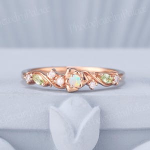 May include: Rose gold ring with a floral design. The ring features a central opal stone, surrounded by smaller gemstones and delicate leaf accents. The ring is set on a light blue surface.