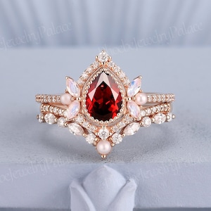 Puede incluir: Un anillo de oro rosa con una piedra roja en forma de pera, adornado con un delicado diseño floral y pequeñas piedras blancas. El anillo está adornado con perlas.