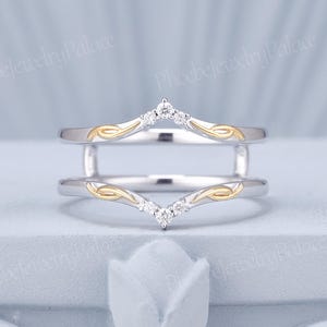May include: A two-piece ring guard featuring a chevron-shaped design in silver-toned metal with yellow gold-toned accents. Each band is adorned with small, clear, round-cut stones at the center point and delicate, swirling metal filigree details.