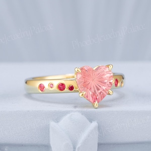 May include: A gold ring featuring a large, faceted, heart-shaped pink gemstone as the centerpiece. The band is accented with several smaller, round, vibrant pink stones set along the sides. The metal has a polished, reflective gold finish.