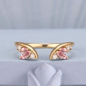 Può includere: Un anello color oro con un design a gambo diviso. Ogni lato presenta un gruppo di pietre preziose rosa e piccole pietre preziose bianche, creando un aspetto a forma di ala. L'anello è posto su uno sfondo azzurro.