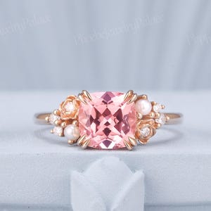 May include: A rose gold ring featuring a large, square-cut pink gemstone as the centerpiece. The ring is adorned with small pearls and accent stones, with floral-inspired detailing on the sides. The ring is set on a light blue surface.