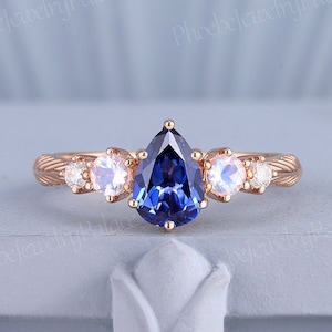 May include: A pear-shaped blue sapphire engagement ring set in rose gold with two smaller moonstone accent stones on each side.