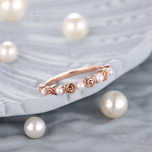 Pode incluir: Um delicado anel de banda de ouro rosa com uma fila de pequenas p&eacute;rolas brancas e detalhes de rosas.