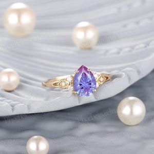 K&ouml;nnte beinhalten: Ein Ring mit einem birnenf&ouml;rmigen, lila-blauen Edelstein, der in eine goldene Fassung gefasst ist und an den Seiten mit kleineren wei&szlig;en Edelsteinen besetzt ist.