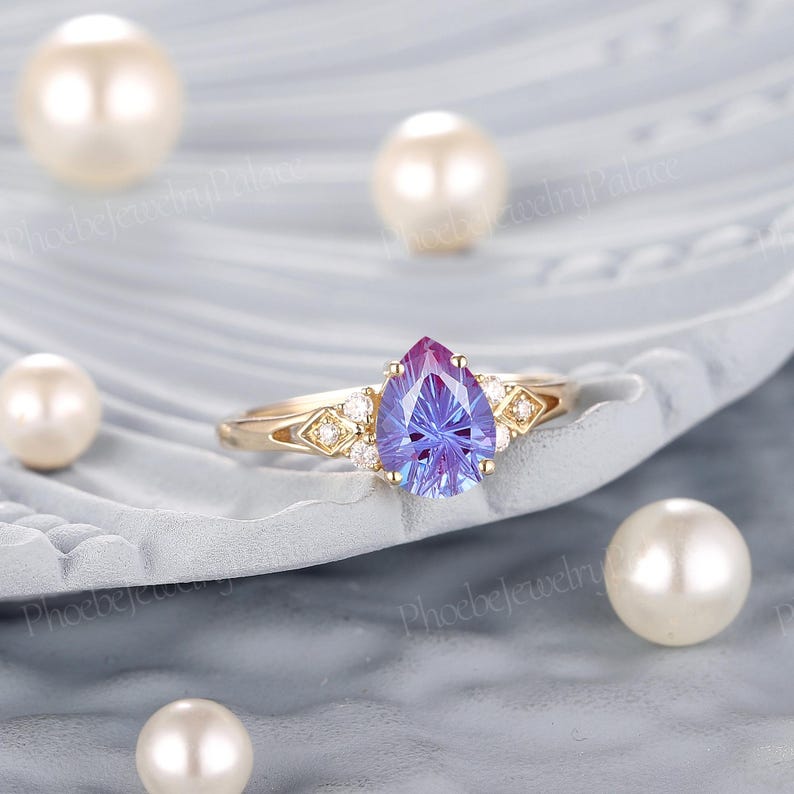 K&ouml;nnte beinhalten: Ein Ring mit einem birnenf&ouml;rmigen, lila-blauen Edelstein, der in eine goldene Fassung gefasst ist und an den Seiten mit kleineren wei&szlig;en Edelsteinen besetzt ist.