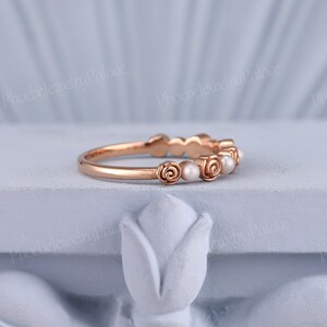 Pode incluir: Um delicado anel de banda de ouro rosa com uma fileira de pequenas p&eacute;rolas e detalhes de rosas.
