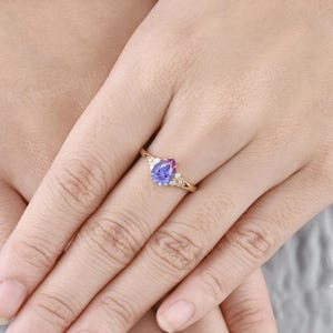 K&ouml;nnte beinhalten: Ein Ring mit einem birnenf&ouml;rmigen, lila-blauen Edelstein, der in Gelbgold gefasst ist und kleinere, klare Edelsteine auf der Ringschiene hat.