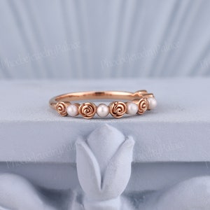 Pode incluir: Um delicado anel de banda de ouro rosa com uma fila de pequenas p&eacute;rolas brancas e acentos de rosas.