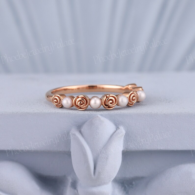 Pode incluir: Um delicado anel de banda de ouro rosa com uma fila de pequenas p&eacute;rolas brancas e acentos de rosas.