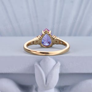 K&ouml;nnte beinhalten: Ein Ring mit einem birnenf&ouml;rmigen, lila und rosafarbenen Edelstein, einem goldenen Band und kleinen Akzent-Steinen. Der Ring liegt auf einem wei&szlig;en Hintergrund.