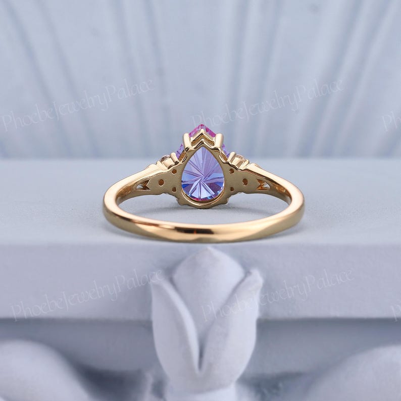 K&ouml;nnte beinhalten: Ein Ring mit einem birnenf&ouml;rmigen, lila und rosafarbenen Edelstein, einem goldenen Band und kleinen Akzent-Steinen. Der Ring liegt auf einem wei&szlig;en Hintergrund.