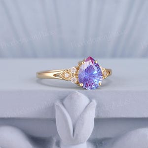 K&ouml;nnte beinhalten: Ein Ring mit einem birnenf&ouml;rmigen Edelstein, der in der Mitte violett und blau ist und von kleineren, klaren Steinen umgeben ist. Der Ring ist in Gelbgold gefasst.