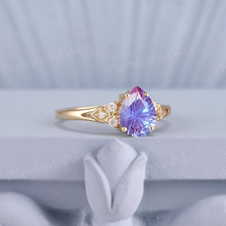 K&ouml;nnte beinhalten: Ein Ring mit einem birnenf&ouml;rmigen Edelstein, der in der Mitte violett und blau ist und von kleineren, klaren Steinen umgeben ist. Der Ring ist in Gelbgold gefasst.