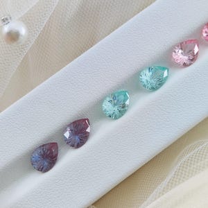 K&ouml;nnte beinhalten: F&uuml;nf birnenf&ouml;rmige Edelsteine in einem Farbverlauf von Rosa bis Blau, auf einer wei&szlig;en Oberfl&auml;che ausgestellt.