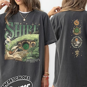 May include: A charcoal gray t-shirt featuring a front graphic of a green hobbit hole, a pipe, and an open book with the text The Shire. The back of the shirt displays a vertical arrangement of four circular icons including a ring and an eye.