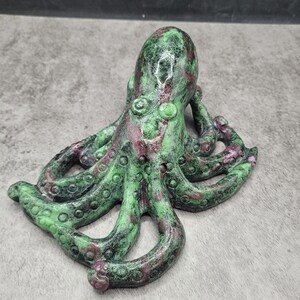 May include: A green and maroon octopus sculpture. The sculpture has a textured surface with a mix of green and maroon colors. The octopus has eight tentacles with suction cups and a rounded head. The sculpture is on a gray surface.