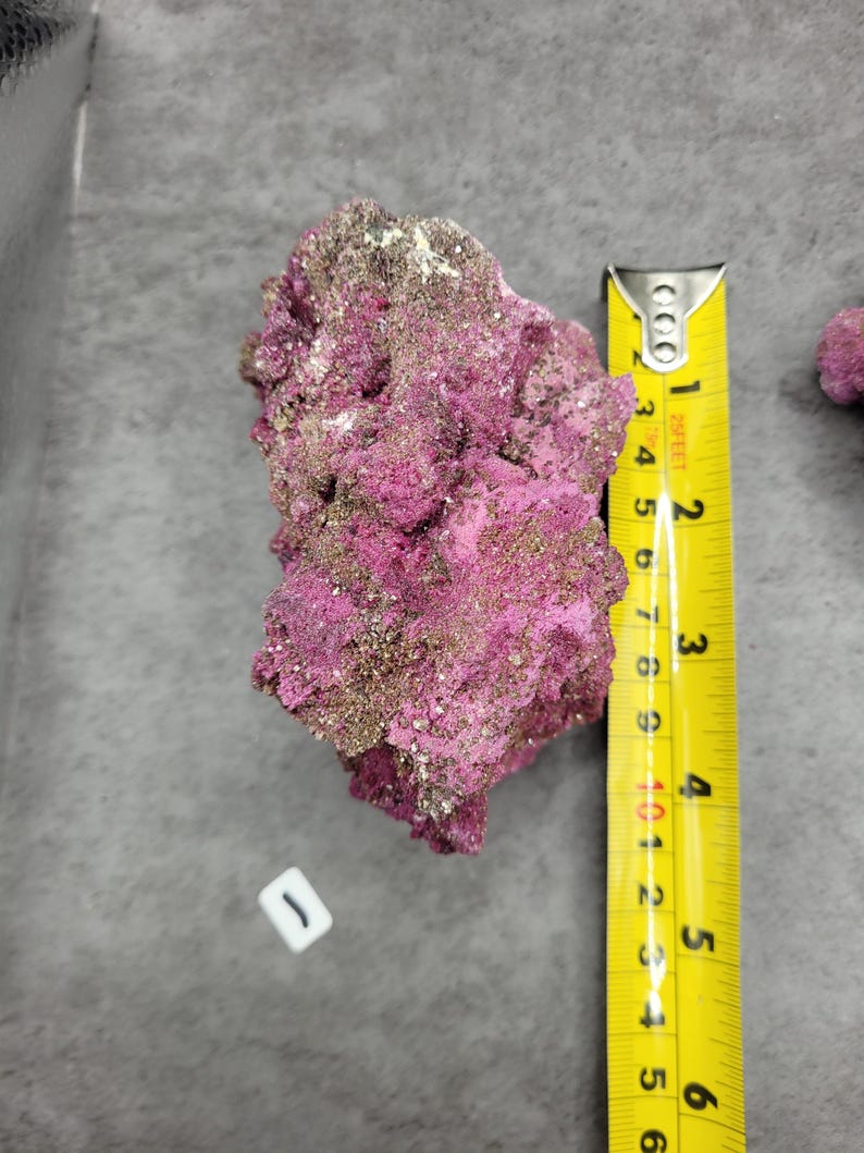 May include: A vibrant pink mineral specimen with a metallic sheen, measuring approximately 5 inches in length. The rock has a textured, porous surface with areas of gold and white. A yellow measuring tape is placed next to the mineral.