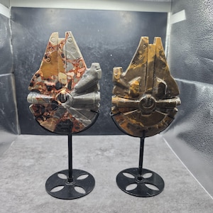 Puede incluir: Dos esculturas de piedra tallada con forma de nave espacial Millennium Falcon, cada una montada sobre un soporte de metal negro. La escultura de la izquierda presenta un patrón moteado marrón, gris y blanco, mientras la derecha es de tono marrón dorado.
