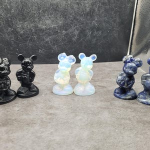 May include: A collection of small Mickey Mouse figurines in various materials. The figurines are in black, opalescent white, and blue. The figurines are standing on a gray surface.