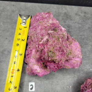 May include: A vibrant pink mineral specimen with a rough, textured surface. The specimen is approximately 3 inches long, as measured by a yellow tape measure. The mineral has patches of green and is set against a gray background.