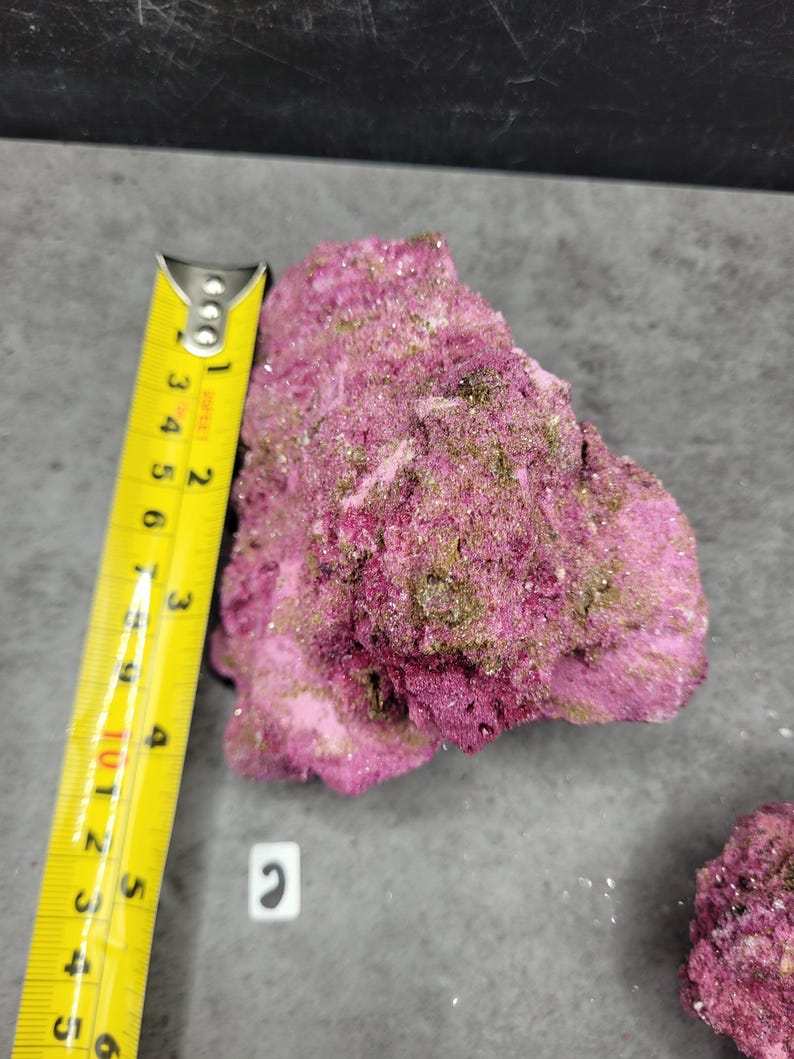 May include: A vibrant pink mineral specimen with a rough, textured surface. The specimen is approximately 3 inches long, as measured by a yellow tape measure. The mineral has patches of green and is set against a gray background.