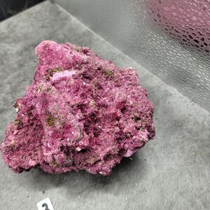 May include: A close-up of a rough, textured rock formation. The rock is a deep magenta color with patches of green and white. A small white tag with the number "3" is visible.