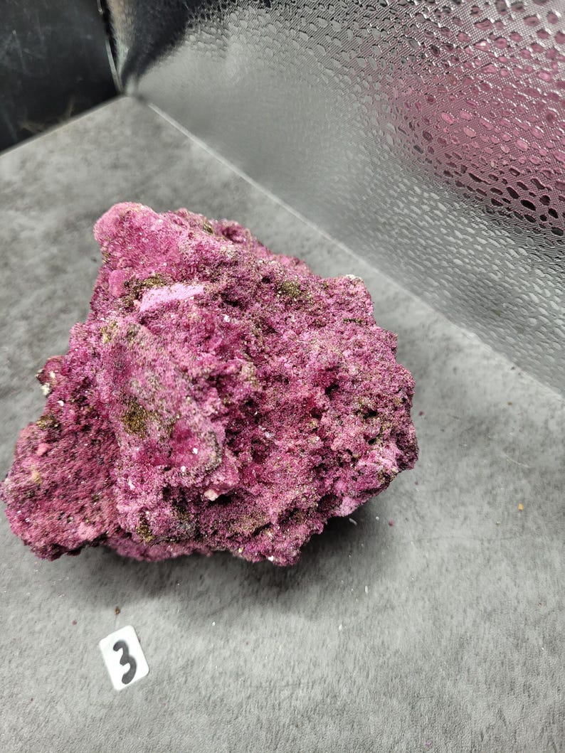 May include: A close-up of a rough, textured rock formation. The rock is a deep magenta color with patches of green and white. A small white tag with the number "3" is visible.