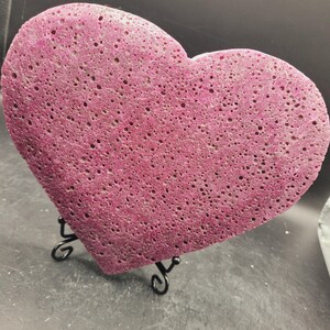 May include: A heart-shaped piece of pink lava rock with a textured surface. The rock is displayed on a black metal stand.