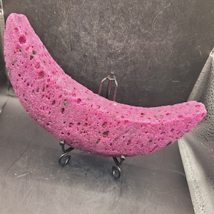 May include: A pink, crescent-shaped sponge with a rough texture, displayed on a black metal stand.