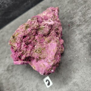 May include: A close-up of a rough, textured rock formation. The rock is primarily a vibrant magenta color, with patches of a muted, greenish-gold hue. The surface appears uneven and crystalline. The rock sits on a gray surface.