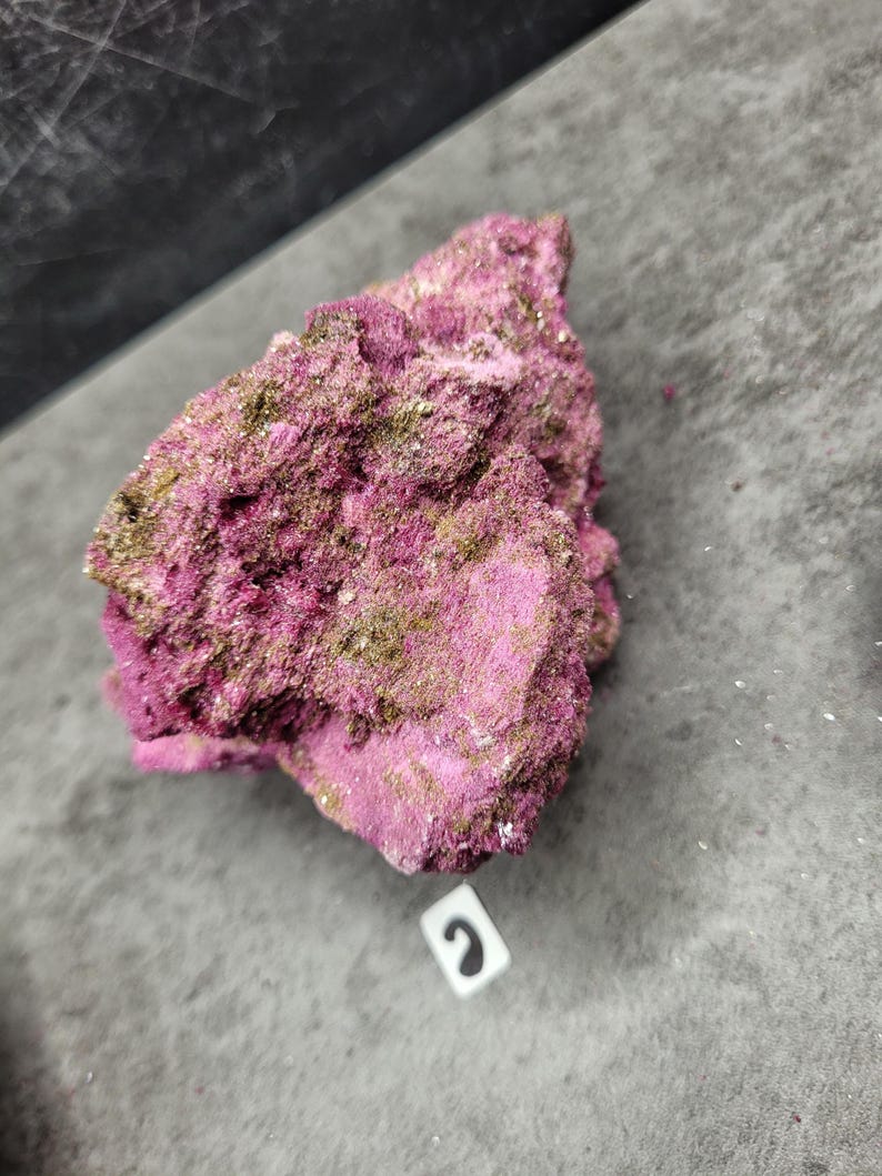 May include: A close-up of a rough, textured rock formation. The rock is primarily a vibrant magenta color, with patches of a muted, greenish-gold hue. The surface appears uneven and crystalline. The rock sits on a gray surface.