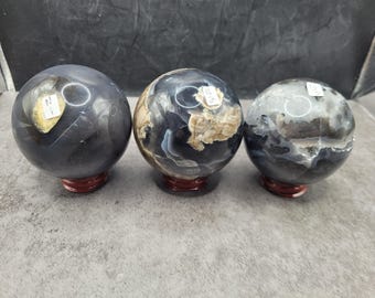 Peut inclure: Trois sphères de pierre polie dans les tons de gris, noir et beige, chacune montée sur un petit socle rond en bois rouge. Les sphères présentent des motifs et des textures uniques, dont certains marbrés.