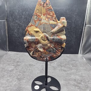 Puede incluir: Una escultura de piedra tallada con forma de nave espacial, que presenta un patrón moteado de colores rojo, marrón, gris y tostado. La escultura está montada sobre un soporte de metal negro con una base circular que tiene cuatro recortes.
