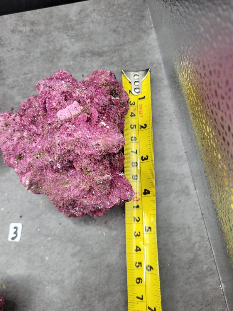 May include: A vibrant, textured, pinkish-purple rock formation. A yellow tape measure shows the rock is approximately 6 inches long. The rock has a rough, uneven surface with small, light-colored inclusions. The number "3" is visible on a small white label.