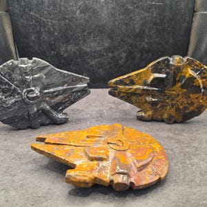 Peut inclure: Trois sculptures en pierre sculptée en forme de vaisseau spatial Millennium Falcon. Une sculpture est gris foncé avec des veines gris clair, tandis que les deux autres sont composées de pierre jaune, orange et brune avec une finition polie.