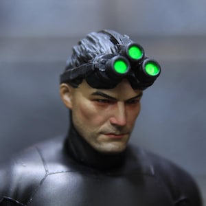 Op de afbeelding: Een zwart actiefiguur met een zwart en groene nachtzichtbril-headset. De figuur heeft kort donker haar en een serieuze uitdrukking.