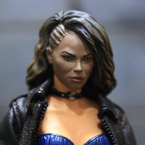 May include: A detailed figurine of a woman with dark skin, styled with a side-shaved hairstyle and wavy brown hair. She wears a black leather jacket, a blue bustier, and a black choker. The figure has a serious expression.