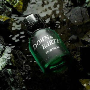 May include: A dark green perfume bottle with the text "DOWN to EARTH MYKONOS" in white. The bottle is partially covered in water droplets and sits among dark rocks and water.