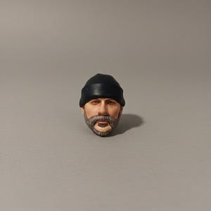 May include: A detailed miniature head of a man with a black beanie, a gray beard, and a neutral expression. The head is painted with realistic skin tones and facial features, showcasing a high level of craftsmanship.