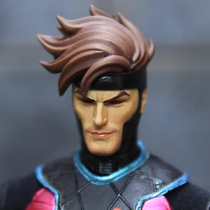 Pode incluir: Um close-up de uma figura de ação do Gambit. A figura tem cabelo castanho, uma faixa preta e uma expressão séria. A fantasia é preta com detalhes em rosa e azul. Os olhos são vermelhos.